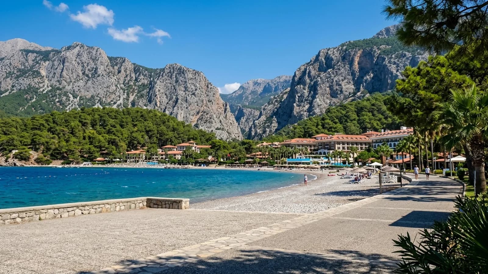 Pebble shore at Göynük with beach hotels among pines and a green valley opening towards grey cliffs.