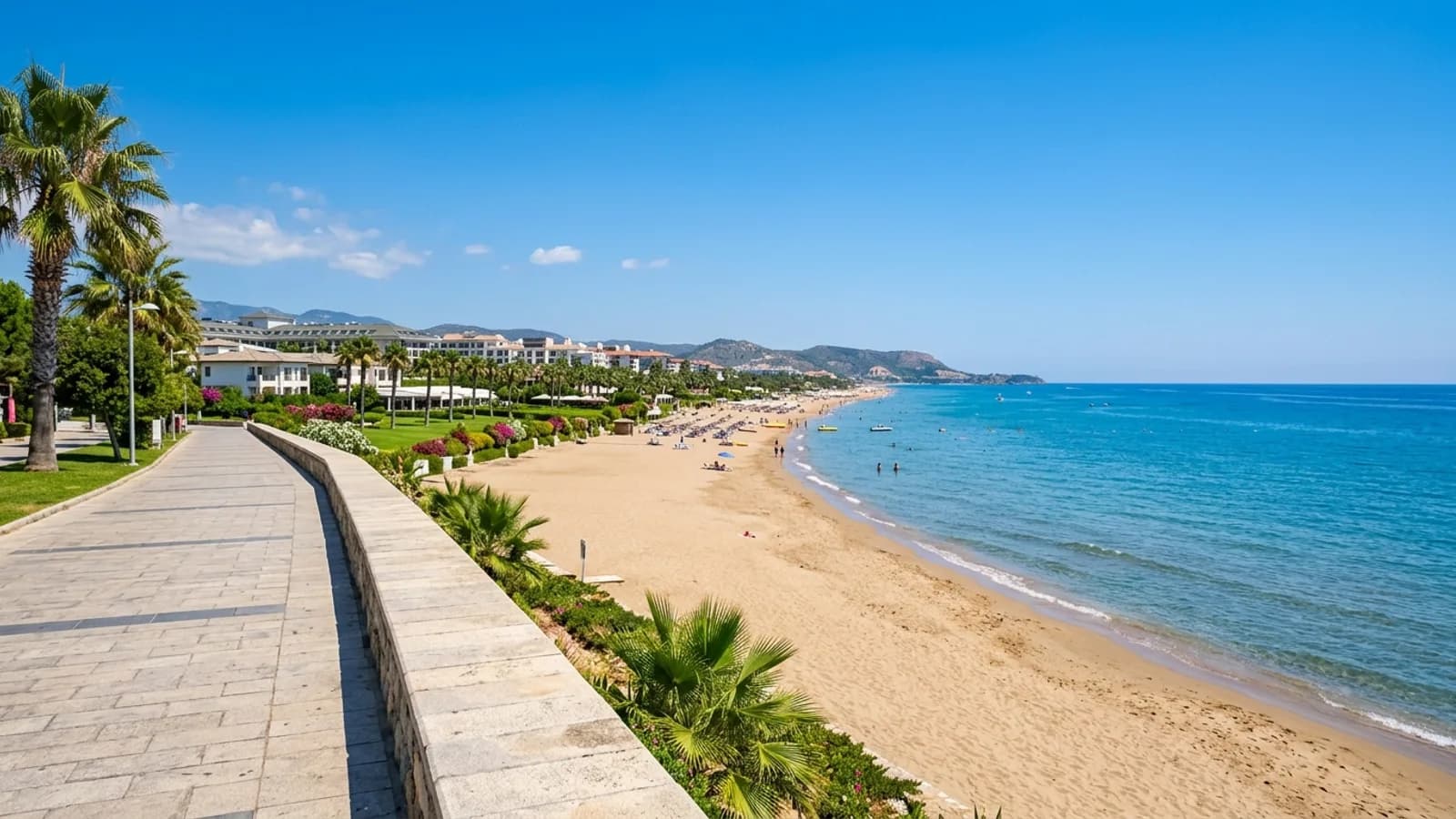 Een lang zandstrand in Gündoğdu met grote resorthotels, groene gazons en een vlakke, kalme zee.