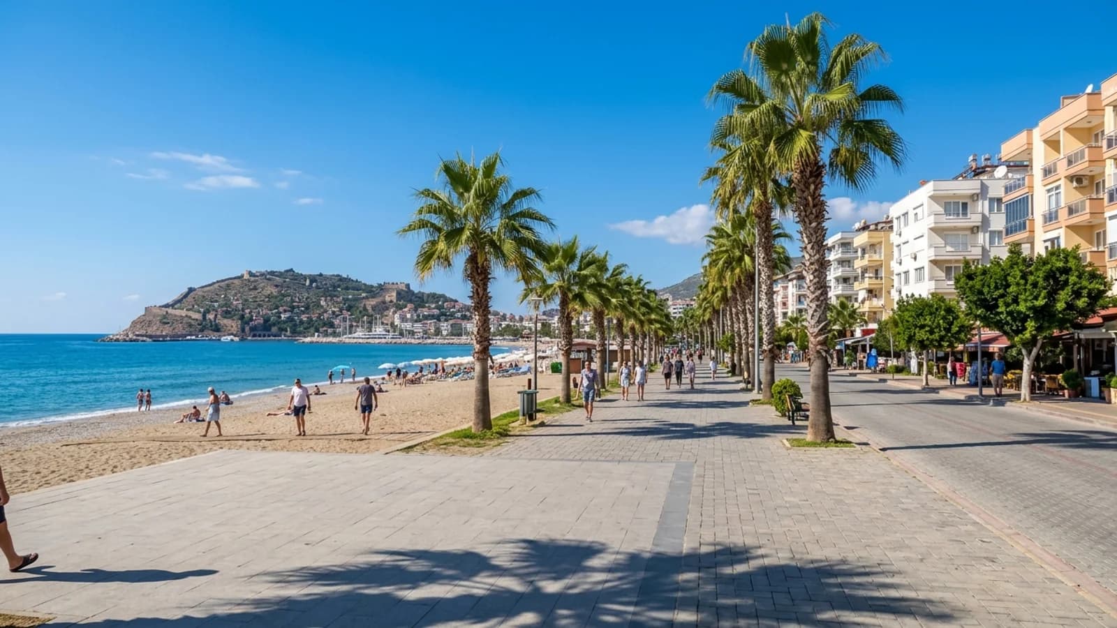 Alanya Oba sahili ve kordon boyu Oba, Alanya'nın palmiyeli sahil yolu ve plajı, arka planda oteller ve uzakta kale yarımadası.
